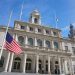 Fachada del Ayuntamiento de Nueva York con la bandera de EE. UU. a media asta en un día soleado.