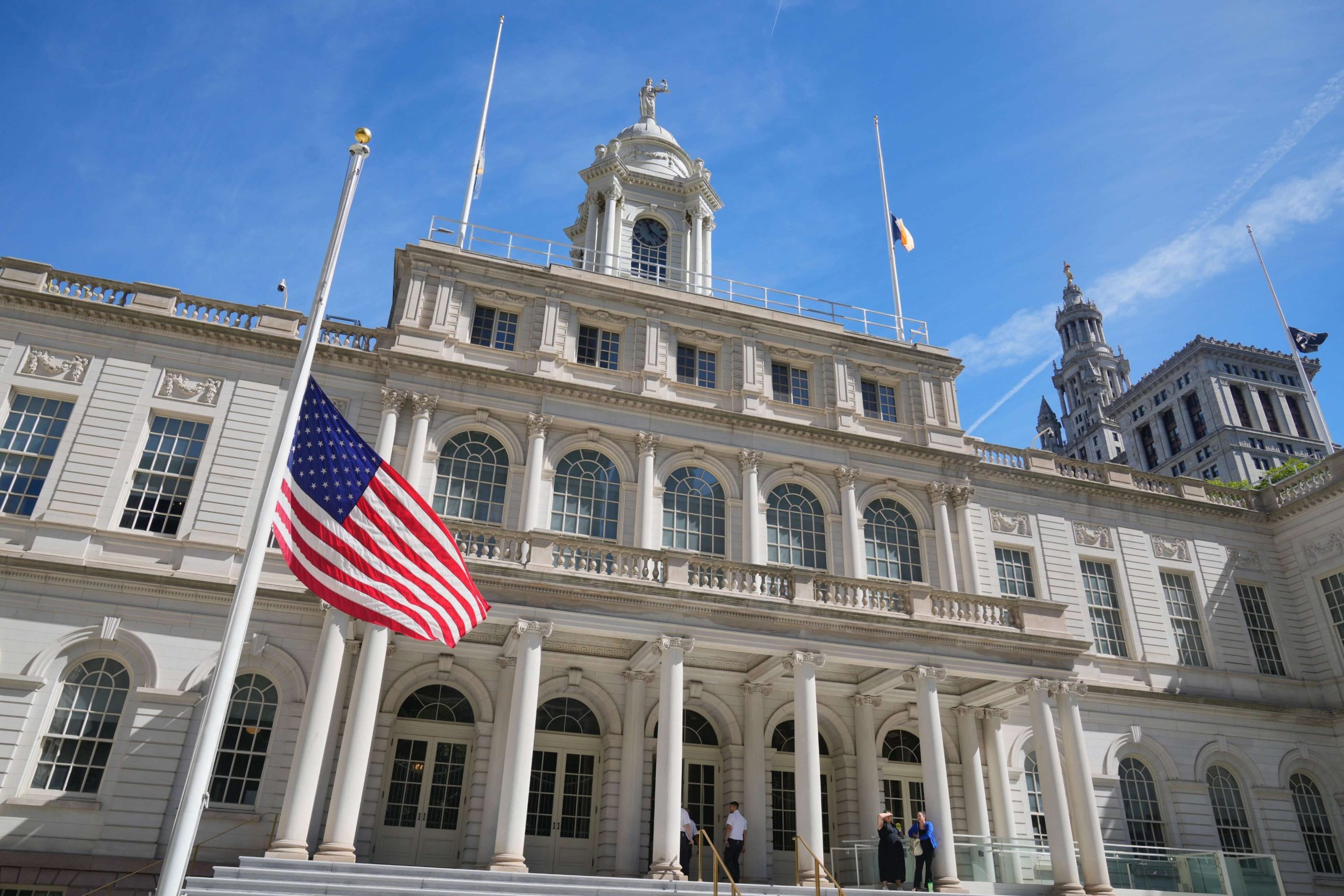 Fachada del Ayuntamiento de Nueva York con la bandera de EE. UU. a media asta en un día soleado.